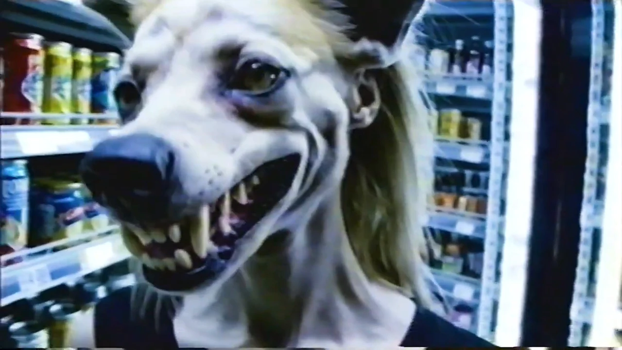 A long-haired subject with an elongated canine-like muzzle and exposed teeth beside a glass-fronted refrigerated display.