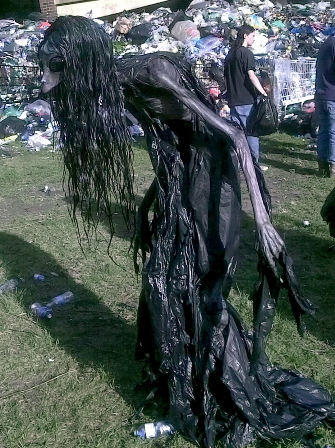A slender trash collector with long dark hair, large black eyes and elongated arms carries a refuse bag near a large pile of rubbish and nearby people.