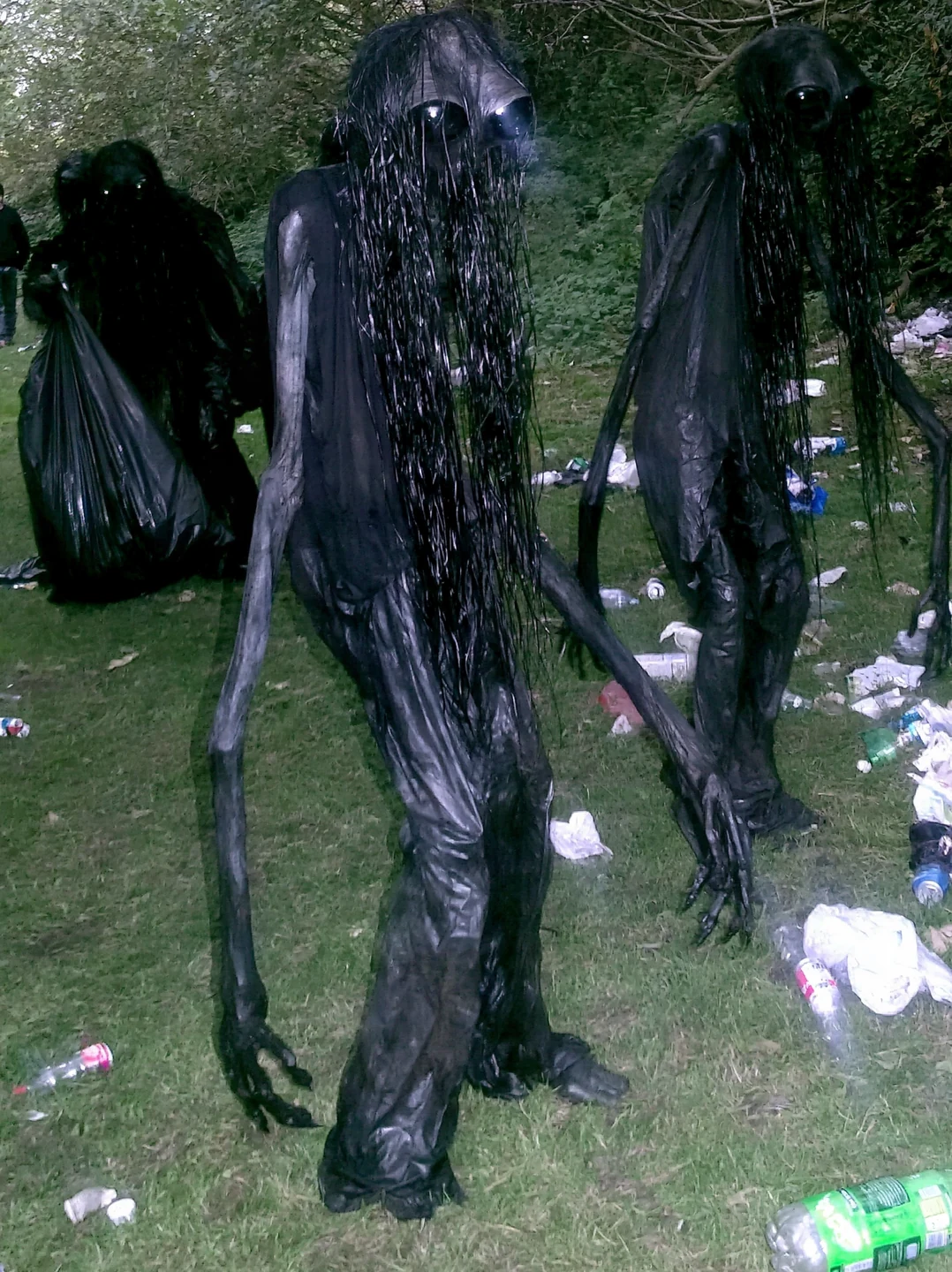 A group of long-haired humanoid trash collectors on littered grass beside trees, with a black refuse bag carried by a figure in the background.